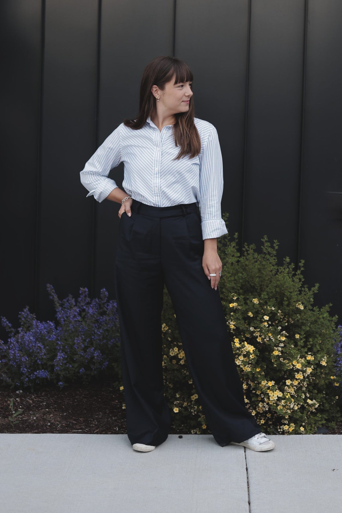 A woman in a striped shirt and navy wool trousers against a black wall with blue and yellow flowers