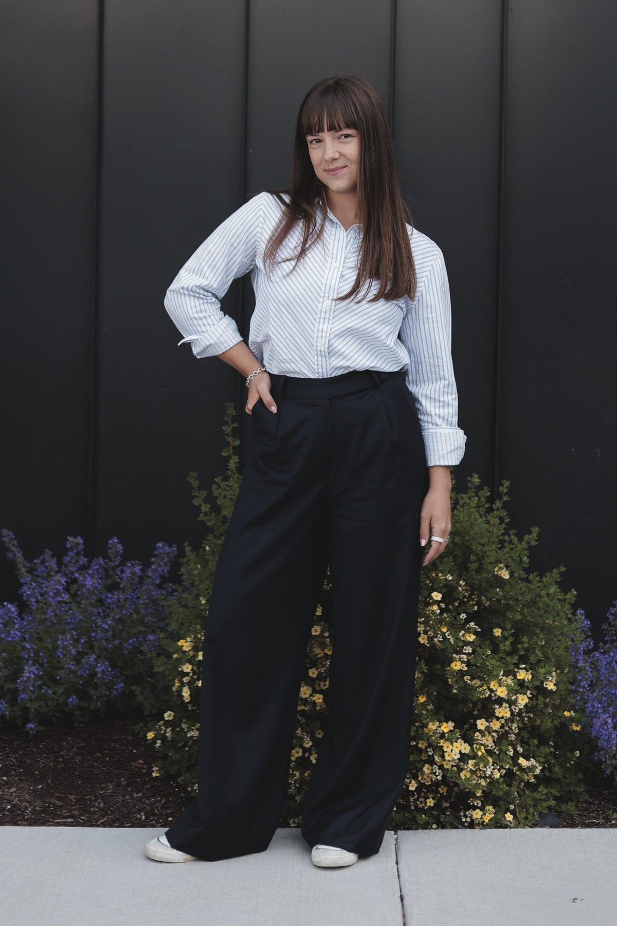 A smiling woman in a striped shirt and navy wool trousers, hand in her pocket